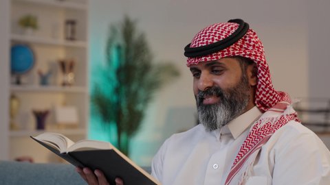 الاهتمام بتصفح الكتب المفيدة, تصويرمقرب لرجل عربي خليجي سعودي يرتدى الثوب والشماغ يجلس على الاريكة يمسك بيده الكتاب, قضاء أوقات الفراغ بالتثقيف والتعلم, حب المطالعة والقراءة, مفهوم التأمل والتفكي