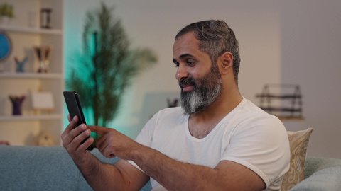 تعابير السرور والاندماج, العمل عن بعد، تصفح التطبيقات وبرامج التواصل الاجتماعي، دمج التقنية والتكنولوجيا في الحياة اليومية، تصوير مقرب لرجل عربي خليجي سعودي يرتدي السروال والفانيله البيضاء يحمل بيده الهاتف المحمول