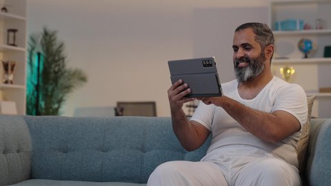 Chatting with family and friends, using tablet apps and programs, integrating modern technology into daily life, a photograph of a Saudi Arabian Gulf man wearing trousers and a white t-shirt holding a tablet, browsing social media.