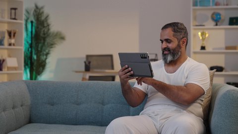 التواصل والدردشة مع الاقارب, تصفح مواقع التواصل الاجتماعي, استخدام تطبيقات وبرامج الأجهزة اللوحية، دمج التكنولوجيا والتقنيات الحديثة فى الحياه اليوميه ، تصويرلرجل عربي خليجي سعودي يرتدي السروال والفانيله البيضاء يحمل بيده جهاز التابلت