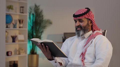 تصوير مقرب لرجل عربي خليجي سعودي يرتدى الثوب والشماغ يجلس على الاريكة يمسك بيده الكتاب, قضاء أوقات الفراغ بالتثقيف والتعلم, حب المطالعة والقراءة, مفهوم التأمل والتفكير, الاهتمام بتصفح الكتب المفيدة