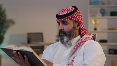A love of reading, the concept of contemplation and reflection, spending leisure time educating and learning, developing reading skills, an interest in browsing useful books, a photograph of a Saudi Arabian Gulf Arab man wearing a thobe and shemagh, sitting on a sofa holding a book.