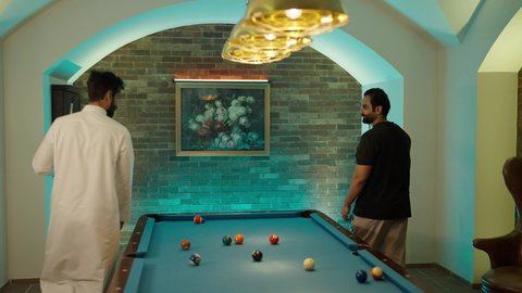 مفهوم المنافسات والتحديات، قضاء أوقات الفراغ مع الاصدقاء, أنشطه وهوايات شبابيه, طاولة بلياردو (السنوكر), شابان عربيان خليجيان سعوديان يرتديان ملابس كاجوال والزي التقليدي يلعبان لعبة البلياردو، صاله رياضيه شبابيه، ايماءات التحية و السلام