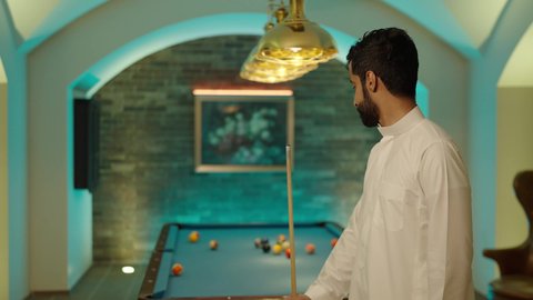 The concept of competitions and challenges, youth activities and hobbies, a youth sports hall, enjoyable leisure time, a billiard table (snooker), an Arab Gulf Saudi young man wearing a white thobe practicing billiards, looking at the camera with expressions of joy.