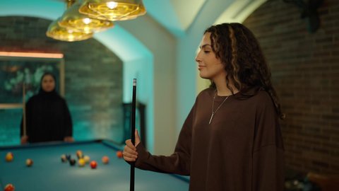 النظر الى الكاميرا بتعابير السعادة و السرور، أنشطة وهوايات شبابية، التسلية والترفيه وقضاء الوقت الممتع, الاستمتاع والشغف بممارسة لعبة السنوكر, تصوير مقرب لشابة عربية خليجية سعودية ترتدي الزي الكاجوال  تمسك بيديها عصا لعب البلياردو