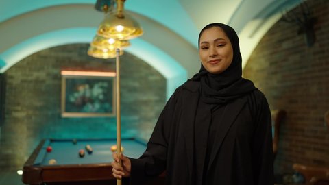 ايماءات السرور والسعادة، مقهى البلياردو, أنشطه وهوايات شبابيه, طاولة بلياردو (السنوكر), امراه عربيه خليجيه سعودية محجبه ترتدي العباءة تمسك مضرب لعبة البلياردو، صاله رياضيه شبابيه