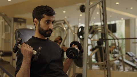 بناء الكتلة العضلية للجسم، استخدام الاثقال الرياضية، رياضة كمال الأجسام، شاب عربي خليجي سعودي يرتدي الزي الرياضي يقوم بممارسة تمارين رفع الأوزان  يحمل بيده الدامبلز، الاهتمام بالصحة واللياقة الجسدية، إيماءات القوة والعزم