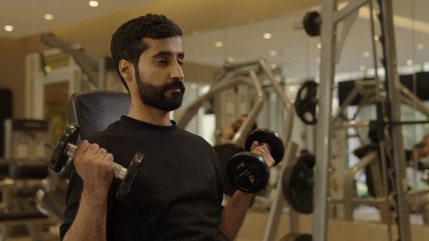 استخدام الاثقال الرياضية، رياضة كمال الأجسام، شاب عربي خليجي سعودي يرتدي الزي الرياضي يقوم بممارسة تمارين رفع الأوزان  يحمل بيده الدامبلز، الاهتمام بالصحة واللياقة الجسدية، إيماءات القوة والعزم، بناء الكتلة العضلية للجسم