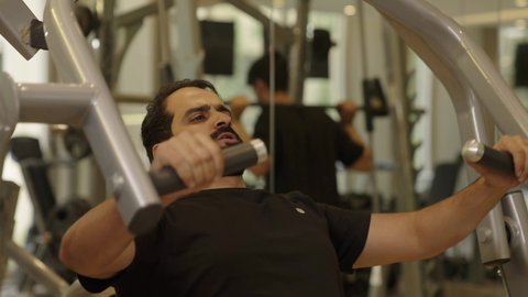 إيماءات القوة والثقة، رياضة كمال الأجسام، أسلوب حياة صحي، الاهتمام بالصحة واللياقة الجسدية، شاب عربي خليجي سعودي يرتدي الزي الرياضي يقوم  باستخدام الأجهزة الرياضية، رجل سعودي يمارس أنشطة لتقوية العضلات