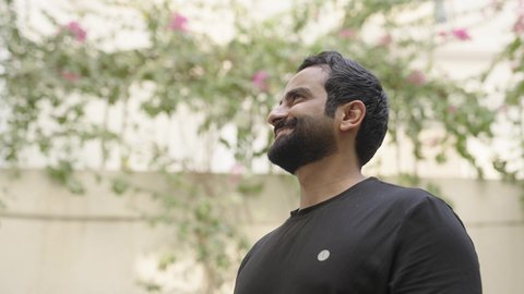 Spending enjoyable time outdoors, in parks and public gardens, an Arab Gulf Saudi man dressed in casual attire is walking in one of the gardens and looking up, enjoying the fresh air and flowers.