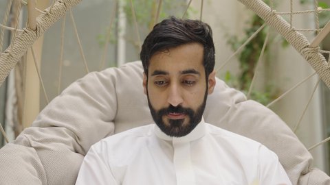 Spending enjoyable time outdoors in one of the tourist chalets, spending free time reading books and novels, a close-up shot of a young Arab Gulf Saudi man wearing a white thobe sitting on a swing reading a book.
