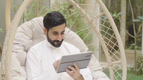 مفهوم التكنولوجيا وإستخدام تقنيات حديثة ، تصوير مقرب لشاب عربي خليجي سعودي يرتدي الثوب الابيض يجلس فى الهواء الطلق على الارجوحة يقوم باستخدام الجهاز اللوحي، ايماءات الاندماج و التركيز ، استخدم جهاز التابلت لانجاز المشاريع والمهام