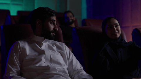 عطلة نهاية الأسبوع، مشاهدة الأفلام والعروض المسرحية في السينما، زوجان عربيان خليجيان سعوديان يرتديان الزي التقليدي يجلسان في صالة السينما، تعابير تدل على السعادة، أنشطة ترفيهية، قضاء أوقات ممتعة