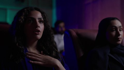 متابعة الفيلم المفضل، الاستمتاع بمشاهدة الأفلام داخل السينما، تعابير الاندماج والتفاعل و الدهشه، امرأة عربية خليجية سعودية محجبة ترتدي عباءه مع صديقتها ، التسلية والترفيه بقضاء أوقات ممتعة
