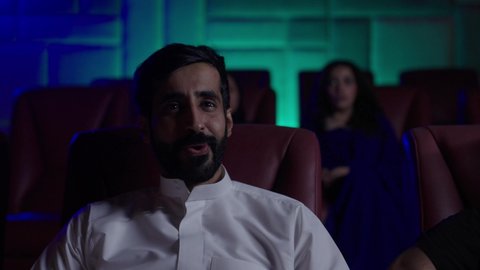 A Saudi Arabian Gulf Arab man wearing a white thobe is sitting in a cinema hall, following the film with gestures of engagement and focus. The modern screening venues in the Kingdom of Saudi Arabia offer watching movies, plays, and various shows, with large screens to enhance the enjoyment of the experience, engaging in fun recreational activities.