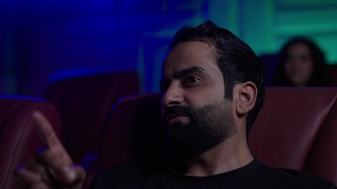 Modern cinemas in the Kingdom of Saudi Arabia, referring to something, watching movies, plays, and various shows, large screens to enhance the enjoyment of the experience, engaging in fun recreational activities, an Arab Gulf Saudi man dressed in casual attire sitting in a cinema hall watching the movie with gestures of immersion and focus.