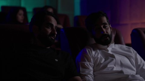 Watching movies, plays, and various shows, large screens to enhance the enjoyment of the experience, engaging in fun recreational activities, two Saudi Arabian Gulf men sitting in a cinema hall watching the movie with gestures of happiness, modern theaters in the Kingdom of Saudi Arabia.