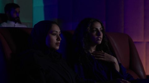 متابعة الفيلم المفضل، الاستمتاع بمشاهدة الأفلام داخل السينما، تعابير الاندماج والتفاعل ، امرأة عربية خليجية سعودية محجبة ترتدي العباءه تجلس برفقة صديقتها، التسلية والترفيه بقضاء أوقات ممتعة