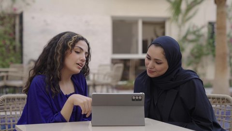 النقر على شاشة الجهاز اللوحي، إجراء مكالمات الفيديو و البث المباشر، تصفح مواقع التواصل الاجتماعي، متابعة الأفلام والمسلسلات المفضلة عبر الإنترنت، امراة عربيه خليجيه سعودية ترتدي العباءة و الحجاب تجلس مع صديقتها تقوم باستخدام جهاز التابلت المحمول