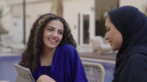 Making video calls and live streaming, browsing social media, watching favorite movies and series online, tapping on the tablet screen, an Arab Gulf Saudi woman wearing an abaya and hijab sitting with her friend using a mobile tablet device.