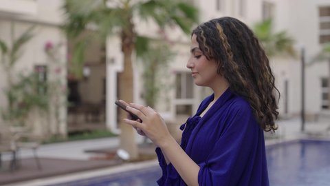 التواصل مع العائلة والأصدقاء، الاستمتاع بالوقت في الهواء الطلق في أحد الشاليهات السياحية، تصوير لشابة عربيه خليجيه سعودية ترتدي الفستان الازرق تقف بجانب حمام السباحة تمسك بيديها الهاتف المحمول
