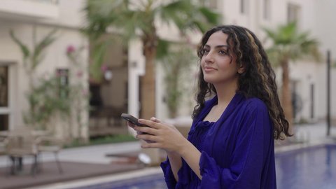 Enjoying time outdoors in one of the tourist chalets, a photo of a young Arab Gulf Saudi woman wearing a blue dress standing by the swimming pool holding a mobile phone in her hands, connecting with family and friends.