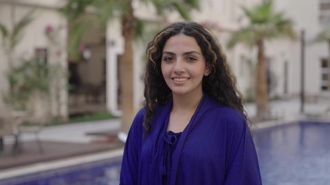 الاهتمام بالأناقة والمظهر الخارجي، مفهوم الأنوثة والنعومة, تصوير مقرب لشابةعربيه خليجيه سعودية ترتدي فستان ازرق تقف بجانب حمام السباحة وتنظر إلى الكاميرا بإيماءات السعادة والسرور, قضاء وقت ممتع فى الخارج