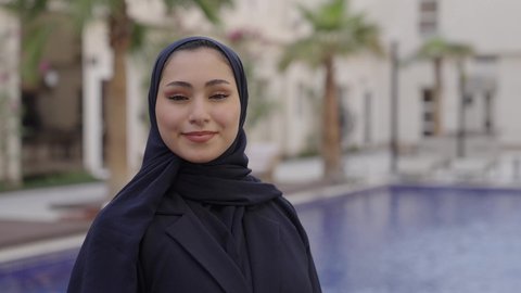 مفهوم الأنوثة والنعومة, الاهتمام بالأناقة والمظهر الخارجي, تصوير مقرب لامراه عربيه خليجيه سعودية محجبه ترتدي العباءة السوداء تقف بجانب حمام السباحة وتنظر إلى الكاميرا بإيماءات السعادة والسرور, قضاء وقت ممتع فى الخارج