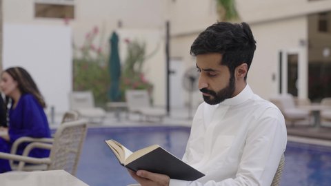 Spending enjoyable time with oneself, the concept of self-learning and self-cultivation, a close-up image of a Saudi Arabian Gulf man wearing traditional attire sitting outdoors reading a book, developing vocabulary.
