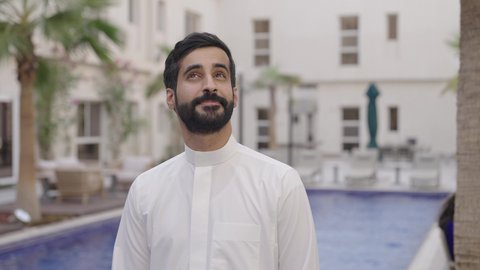 الاستمتاع بالهواء الطلق، شاب عربي خليجي سعودي يرتدي الثوب الابيض يقوم بالمشي بجانب حمام السباحة و ينظر الى الاعلي ، قضاء وقت ممتع فى الخارج، ايماءات السعادة و السرور