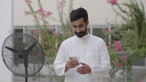 Browsing social media on a mobile phone, using modern technological devices, a stroll outdoors, a young Arab Gulf Saudi man wearing traditional attire holding a mobile phone in his hand, gestures of happiness and joy.