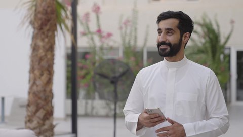 استخدام الأجهزة التقنية الحديثة، نزهه في الهواء الطلق, تصفح وسائل التواصل الاجتماعي عبر الجوال,شاب عربي خليجي سعودي يرتدي الثوب التقليدي يمسك الهاتف المحمول بيده، ايماءات السعادة و السرور