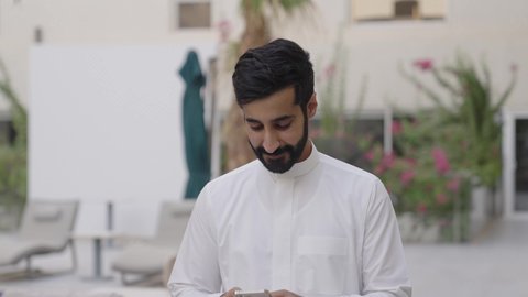 نزهه في الهواء الطلق، استخدام الأجهزة التقنية الحديثة, تصفح وسائل التواصل الاجتماعي عبر الجوال,شاب عربي خليجي سعودي يرتدي الثوب التقليدي يمسك الهاتف المحمول بيده، ايماءات السعادة و السرور
