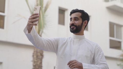 دردشات وحوارات ودية مع الأصحاب، التواصل مع الأصدقاء والأقارب بإيماءات السرور, مفهوم صلة الرحم, شاب عربي خليجي سعودي يرتدي الثوب الابيض يقوم بإجراء مكالمة فيديو عبر الجوال, استخدام تطبيقات التواصل الاجتماعي، الاستمتاع بقضاء عطلة صيفية