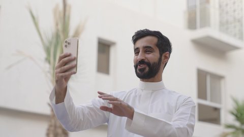 التواصل مع الأصدقاء والأقارب بإيماءات السرور, مفهوم صلة الرحم, شاب عربي خليجي سعودي يرتدي الثوب الابيض يقوم بإجراء مكالمة فيديو عبر الجوال, دردشات وحوارات ودية مع الأصحاب, استخدام تطبيقات التواصل الاجتماعي، الاستمتاع بقضاء عطلة صيفية