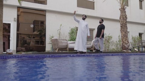 التقاط صورة سيلفي تذكارية باستخدام الهاتف المحمول، اجراء مكالمة فيديو والتواصل مع الاهل والاصدقاء، دمج التقنيات والتكنولوجيا الحديثة بالحياة اليومية،رجل عربي خليجي سعودي يرتدي الثوب الابيض يقف برفقة صديقه بجانب حمام السباحة يمسك بيدية الجوال