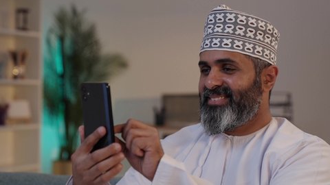 Zooming in and out of the screen, browsing websites, using advanced modern technology in daily life, gestures of happiness and joy, an elderly Arab Gulf Omani man smiling, wearing a dishdasha and a kumma, holding a mobile phone in his hand.