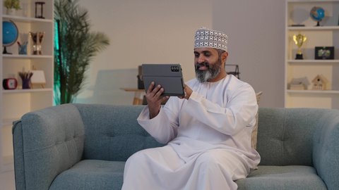 التواصل عبر النت, استخدام الأجهزة و التقنيات الحديثة، مفهوم إدارة المشاريع عن بعد، ايماءات السعادة و السرور، رجل شايب عربي خليجي عماني مبتسم يرتدي الدشداشة و الكمة يمسك بيده التابلت