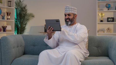 التواصل مع الأهل و الأصدقاء، استخدام الأجهزة و التقنيات الحديثة، مفهوم إدارة المشاريع عن بعد، ايماءات السعادة و السرور، رجل شايب عربي خليجي عماني مبتسم يرتدي الدشداشة و الكمة يمسك بيده التابلت