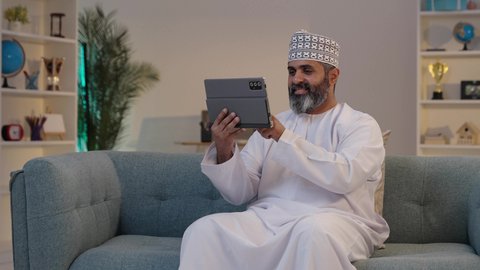 انجاز مهام العمل عبر الجهاز اللوحي، ايماءات السعادة و السرور، استخدام الاجهزة التقنية الحديثة، مفهوم إدارة المشاريع عن بعد، رجل شايب عربي خليجي عماني مبتسم يرتدي الدشداشة و الكمة يمسك بيده التابلت