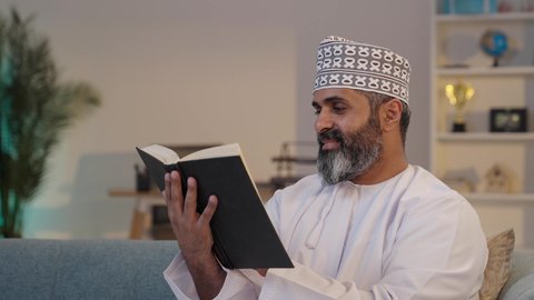 Spending free time learning, reading novels and stories, expressions of focus and enjoyment, a smiling elderly Arab Gulf Omani man wearing a dishdasha and a kumma holding a book in his hand, interest in knowledge and culture.