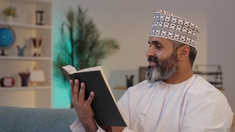 تمضية وقت الفراغ بالتعلم و التثقيف الذاتي، قراءة الرواية والقصة، تعابير التركيز و الاستمتاع،  رجل شايب عربي خليجي عماني مبتسم يرتدي الدشداشة و الكمة يمسك بيده كتاب