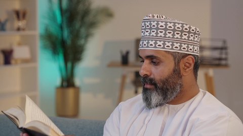 مطالعة الكتب و الروايات، ثقافة التعلم الذاتي, رجل شايب عربي خليجي عماني مبتسم يرتدي الدشداشة و الكمة يمسك بيده كتاب, إيماءات التركيز و الاندماج, قضاء وقت ممتع