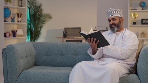 الجلوس على أريكة مريحة, مطالعة الكتب و الروايات، ثقافة التعلم الذاتي, رجل شايب عربي خليجي عماني مبتسم يرتدي الدشداشة و الكمة يمسك بيده كتاب, إيماءات التركيز و الاندماج, قضاء وقت ممتع
