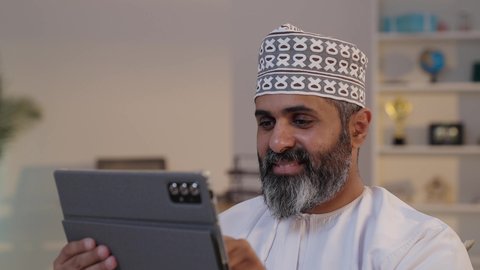 العمل عن بعد من خلال الانترنت، ايماءات السرور والسعادة, رجل شايب عربي خليجي عماني مبتسم يرتدي الدشداشة و الكمة يمسك بيده التابلت, تصفح مواقع التواصل الاجتماعي