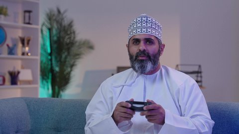 الاستمتاع بألعاب الفيديو, قضاء أوقات الفراغ بلعب البلايستيشن, رجل شايب عربي خليجي عماني مبتسم يرتدي الدشداشة و الكمة يمسك بيده عصا التحكم, ايماءات التركيز و الاندماج, تقنيات حديثة مسلية, أنشطة ترفيهية ممتعة