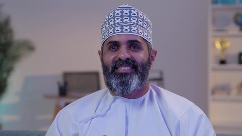 قضاء أوقات الفراغ بلعب البلايستيشن, رجل شايب عربي خليجي عماني مبتسم يرتدي الدشداشة و الكمة يمسك بيده عصا التحكم و يلعب بألعاب الفيديو, ايماءات التركيز و الاندماج, تقنيات حديثة مسلية, أنشطة ترفيهية ممتعة