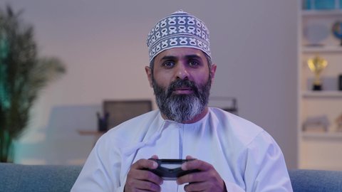 Modern entertaining technologies, enjoyable recreational activities, spending free time playing PlayStation, a smiling elderly Arab Gulf Omani man wearing a dishdasha and kumma holding a game controller and playing video games, gestures of focus and immersion.