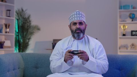 الألعاب الالكترونية و الأجهزة الحديثة، مفهوم الاندماج والتفاعل باللعب ألعاب البلايستيشن، قضاء أوقات ممتعة,  رجل شايب عربي خليجي عماني مبتسم يرتدي الدشداشة و الكمة يمسك بيده عصا التحكم  ايماءات التركيز و التوتر