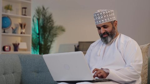 استخدام الكمبيوتر لإرسال البريد الإلكتروني، التواصل مع العملاء عن بعد، دمج التقنيات الحديثة و المتطورة مع العمل, رجل شايب عربي خليجي عماني مبتسم يرتدي الدشداشة و الكمة و يستخدم اللابتوب